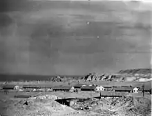 A black and white photograph of a desert landscape; in the mid distance are several huts