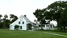 A color photo of a white two-story building with black shutters, a tall wood-shingled roof, and an outdoor veranda. The first floor has a stucco finish and has one window, the second story is wood clapboard and has two windows topped with a chimney. A lawn and trees surround it.