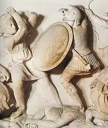 Two figures engaged in combat. The figure on the right is donned with armor and a shield; the one on the left appears to be unprotected and in a weaker position.