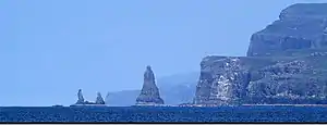 A large sea stack and two smaller ones stand off a coast of high cliffs. There is a deep blue sea in the foreground and two further peninsulas with high cliffs beyond.