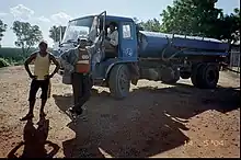 Three men show off a water tanker. Two stand in front of it, one holding the door open, while the third sits at the steering wheel. The water tanker is blue and appears to have "13" written on the side.