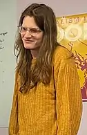 A white woman with brown eyes and long, brown hair. She is wearing clear glasses and a yellow sweater while facing slightly to the left.