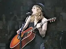 Image of a blond woman. She is dressed all in black but showing her arms, and wearing a hat of the same color. She is playing an orange-red acoustic guitar while a microphone is to her mouth. She is looking to her right and smiling