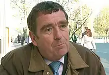 A man in a brown jacket, shirt and tie is pictured sitting outdoors in late spring