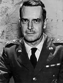 Man with dark hair and moustache in a dress uniform, suit and tie, sitting at a table, with a star indicating his rank, in front of a map on a wall.