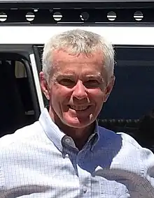 A white, grey-haired man looks at the camera and smiles. He is wearing a collared shirt and standing in sunlight.