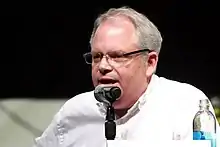 A Caucasian man wearing a white shirt speaks into a microphone.