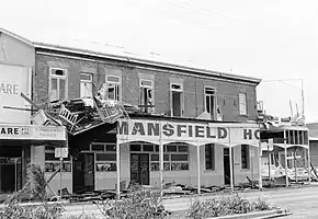 Black and white photograph of storefronts with broken windows, some structural damage, and debris littering the sidewalk