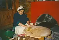 Traditional tableside preparation of gözleme in a restaurant near Antalya. A gas-powered sac griddle can be seen inside the red oven to the right, with two thin wooden rolling pins ("oklava") and pastry brushes used to prepare the dough to the lower left. The cook is seen cutting up the finished gözleme on her pastry board table to prepare for final serving.