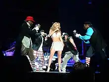 A blonde woman wearing a white top and short skirt sings. She is flanked by four men, who pretend to take photographs of her.