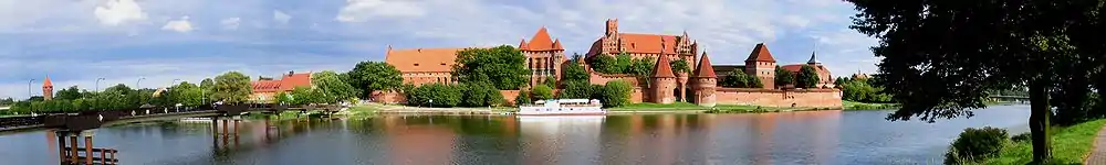 An orange brick castle with a curtain wall and a central keep. The site is surrounded by water. The gateway is flanked by two round towers with high peaked roofs. Aside from the keep, there is another building within the castle rising above the curtain wall.