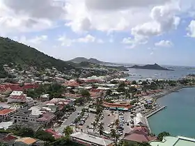 A village on the coast of an island. Small buildings are located throughout the island, with mountains in the background and the ocean on the right. Among the buildings in the foreground is a parking lot adjacent to a marina. A peninsula stretches out into the ocean and boats are on the ocean in the background.