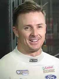 A man in his mid-30 smiling away from the camera. He is wearing a white fireproof vest with sponsors logos.