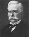 A headshot of a Caucasian male with a white handlebar moustache and white hair. He is wearing a white winged-collar shirt, black cravat and black coat.