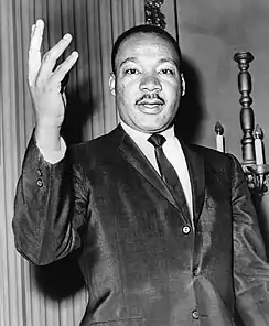 Black-and-white photo of an African-American man, wearing a coat and tie, making an open-handed gesture.