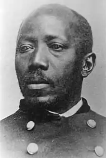 Black and white photo of a Black man of steady countenance and with short, receding hair, and a short goatee. He is wearing a military jacket over a white shirt.