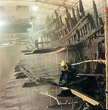 A diagonal section of a wooden ship seen from the stern while being sprayed with a clear liquid from a built-in sprinkler system; on the remnants of the main deck there is a person in a black, full-body protective plastic suit with a yellow helmet