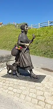 A bronze statue of Mary Anning walking with a pickaxe and fossel in her hand. A dog is walking beside her.