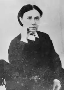 A dark haired woman wearing dark clothing, right hand on chin.