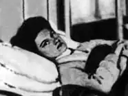 A white woman with dark hair is lying in a hospital bed; she is looking at the camera