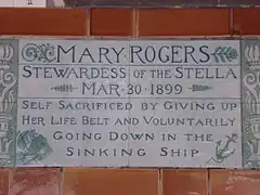 A tablet formed of two large tiles, bordered by green flowers in the style of the Arts and Crafts movement, and decorated with a stylised ship and anchor. The tablet reads "Mary Rogers, Stewardess of the Stella, March 30, 1899, Self sacrificed by giving up her life belt and voluntarily going down in the sinking ship".