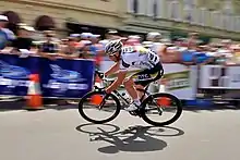 A road racing cyclist in a white and black jersey with green and gold trim. Spectators are visible behind roadside barricades behind him, though they are out of focus.