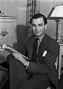 A Caucasian man wearing a suit, sitting on a chair with legs crossed, facing slightly left, and holding a newspaper in his lap