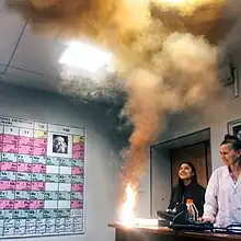 An instructor and a student look at clouds of smoke and a bright flame produced on the edge of a table.