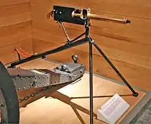 Brass barrelled gun with black coloured rear parts. On a tripod on a wooden plinth with a wood wall behind.