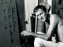 Colin McCahon in his studio, shirtless and holding a cigarette.