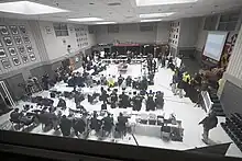 Folding tables are arranged in rows and a square in a large presentation room in a police station. People in various uniforms representing many agencies are seated at the tables and focused on their laptops. The center of the room has a table piled with boxes of pizza, salads, and other provisions.