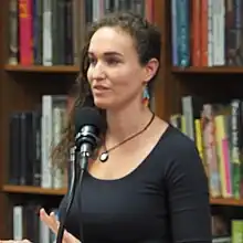 photograph of a woman standing at a microphone, in front of bookshelves