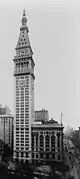 Aerial view of a thin, 50-story building with a light exterior; the building has a pyramidal roof with a large spire, and a clock is visible below the roofline.