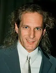 Middle-aged Caucasian man with long, blond hair, smiling while looking directly into the camera. He is wearing a collared shirt, suit and tie.