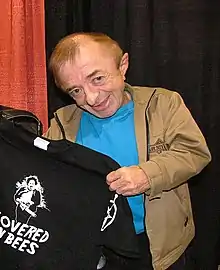 A short man in a jacket holding up a T-shirt to pose for a picture with it