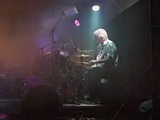 A man with gray hair drums behind his drum-set in a faintly lit room.