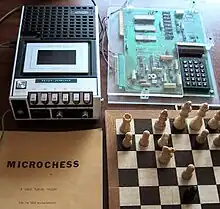 A chess game being played on a board, next to a circuit board with several buttons as inputs connected to a cassette player