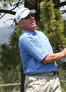 A man in his late fifties wearing a white baseball cap and a light-blue polo shirt looks to the right of the image, following a golf ball he just hit.