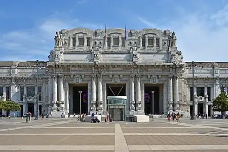 An ornate and extensively detailed gray stone classical portico with four sets of twinned columns at the top of which the Italian and European Union flags hang limply; above is clear blue sky. In front is a more modern one-story pavilion with an elevator in front of it inside a circular wall. Some people can be seen walking around.