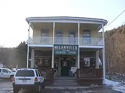 Old-fashioned, refurbished, two-story building, with yellow and blue paint and white siding. A sign on the front of the building reads "MILANVILLE GENERAL STORE, EST. 1850," and there a few cars parked in front on it.