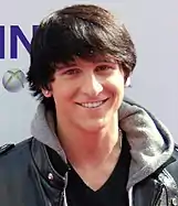 A head shot of a teenage boy with brown, straight hair, wearing a grey, hooded jacket. He is posing at a press event and smiling.