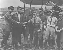 A monochrome photograph of eight variously dressed military airmen standing smiling in front of a biplane, with a tall brigadier general in uniform shaking hands with a short-statured, hatless man wearing a long-sleeved shirt and a tie