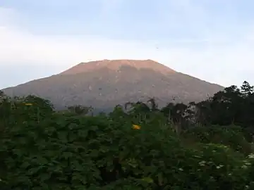 A photograph depicting a blue sky with white clouds at the top, a grey mountain range in the middle, and green foliage at the bottom.