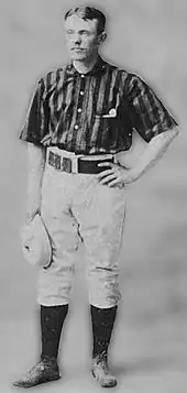 A baseball player standing, facing slightly to his right, with his right arm relaxed and holding a baseball cap, and his left arm raised so that his left hand his resting on his hip