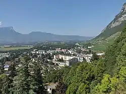 Photo shows homes and apartments in the center and a mountainside to the right. In the background are more mountains.