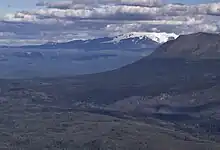 A glaciated, flat-topped, gently-sloping mountain shadowed by clouds with a much lower barren mountain in the right foreground.