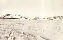 A mountain with a rounded summit and snowy slopes, with smaller hills to its right, rising from flat snow-covered ground