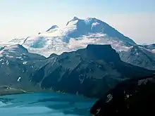 A prominent, glaciated mountain rising over a flat-topped, rocky mountain and a turquoise-coloured alpine lake in the foreground.