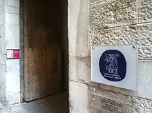 An open door on a stone wall. To the right there is a plastic sign with a round logo.