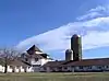 Two wings of a white barn with two silos under a blue sky with cirrus clouds